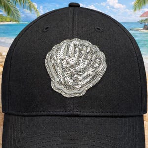 May include: Black baseball cap with a sequined seashell design. The cap has a curved peak and a structured crown. The seashell is silver and has a detailed, textured appearance. The background shows a beach scene with palm trees and blue skies.
