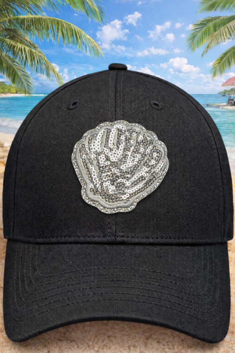 May include: Black baseball cap with a sequined seashell design. The cap has a curved peak and a structured crown. The seashell is silver and has a detailed, textured appearance. The background shows a beach scene with palm trees and blue skies.