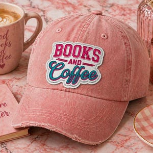 May include: A dusty rose baseball cap with the text "BOOKS AND Coffee" in a white and teal patch. The cap is distressed and has a curved brim. A pink mug with coffee and a book are in the background.