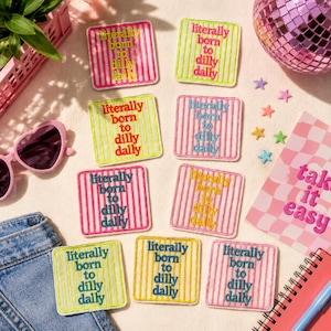 May include: Eight square embroidered patches featuring vertical stripes and the text literally born to dilly dally in various color combinations including pink, yellow, green, and blue, arranged on a flat surface with other accessories.