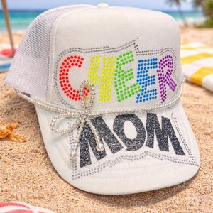 May include: White trucker hat with a mesh back, featuring the word "CHEER" in colorful rhinestones and "MOM" in black glitter. A pearl bow and accents add a decorative touch. The hat is resting on a sandy beach.