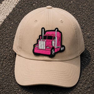 May include: A tan baseball cap featuring a pink semi-truck patch. The cap has a curved brim and a classic six-panel design. The truck patch is outlined in black and has a detailed design.