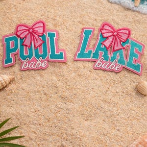 May include: Two pink and teal embroidered patches with the words "POOL babe" and "LAKE babe" are displayed on a sandy beach. Each patch features a pink bow. Seashells, a starfish, and a woven basket are also visible.