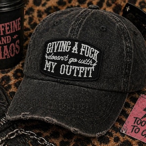 May include: Distressed black baseball cap with the text "GIVING A FUCK doesn't go with MY OUTFIT" on a patch. The cap is on a leopard print surface, with a coffee cup and sunglasses nearby.