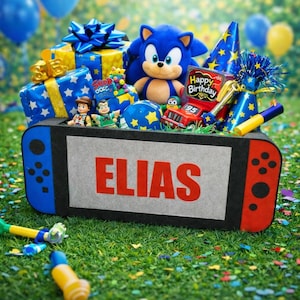 May include: A birthday-themed gift box shaped like a game console, filled with presents, a Sonic the Hedgehog plush, and party favors. The box has the name "ELIAS" in red letters. Blue and yellow balloons and confetti are scattered around.