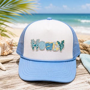 May include: A white and light blue trucker hat featuring a front patch with the word HOWDY spelled out in embroidered ocean-themed letters, including coral, a seashell, blue waves, and seaweed designs, with a mesh back and a solid brim.