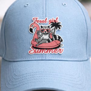 May include: Light blue baseball cap featuring a patch with a raccoon wearing sunglasses, relaxing on a flamingo float with a drink. The patch reads "Feral girl Summer" in pink script. A palm tree is also included in the patch design.