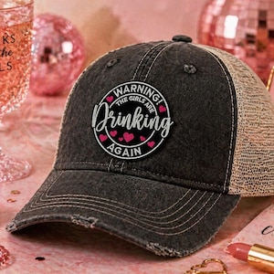 May include: A distressed black trucker hat with a beige mesh back. The hat features a circular patch with the words "WARNING! THE GIRLS ARE DRINKING AGAIN" in white script and pink hearts. The hat is on a pink surface.