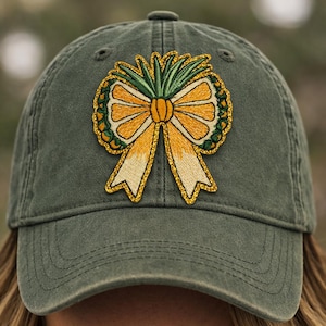 May include: Olive green baseball cap featuring an embroidered design of a yellow and gold bow with green accents. The bow has a pineapple-like design in the center. The cap has a curved brim and a classic six-panel construction.