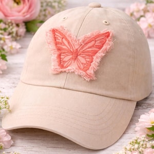May include: A beige baseball cap with a pink embroidered butterfly patch. The butterfly has a fringed edge. The cap has a curved brim and a button on top. The background includes pink flowers.