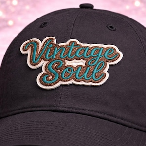May include: Black baseball cap with the embroidered text "Vintage Soul" in teal and brown. The hat has a curved brim and a fabric strap closure. The hat is a classic style.