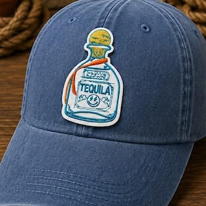May include: Blue baseball cap with a tequila bottle patch. The patch features a white bottle with the word "TEQUILA" and a yellow and orange design. The word "BLUE" is at the bottom.