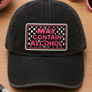 May include: Black baseball cap with a rectangular patch that reads "MAY CONTAIN ALCOHOL" in pink and white lettering. The patch has a pink border and a black and white checkered pattern. The cap is on a wooden surface.