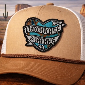 May include: Tan and white trucker hat with a brown brim and a heart-shaped patch. The patch features the words "TURQUOISE & TATTOOS" in white lettering on a turquoise and brown design. The hat has a rope detail.
