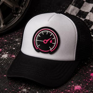 May include: A black and white trucker hat featuring a circular patch on the front panel. The patch displays a black speedometer gauge graphic with a bright pink needle and circular border. The hat has a solid black brim and mesh back panels.