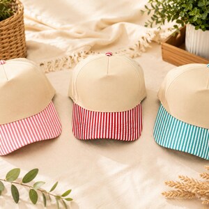 May include: Three baseball caps with solid beige crowns and striped brims. The left cap has a pink and white striped brim, the center cap has a red and white striped brim, and the right cap has a teal and white striped brim. Each cap features a small button.