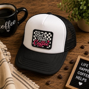 May include: A white and black trucker hat featuring a rectangular patch with a black and white checkered background. The patch displays the text "But First, Coffee" in white and pink script lettering, accompanied by a small pink heart icon.