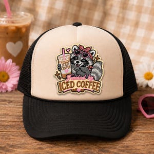 May include: A beige and black trucker hat featuring a graphic of a raccoon wearing a pink patterned bandana and heart-shaped sunglasses while holding a cup of iced coffee. The text "Fueled by ICED COFFEE" is printed in brown and pink below the raccoon.