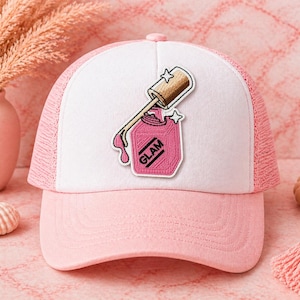May include: A pink and white trucker hat with a pink brim and mesh back. The hat features a patch of a pink nail polish bottle with the word "GLAM" and a dripping design. The nail polish bottle has a gold cap and two star accents.