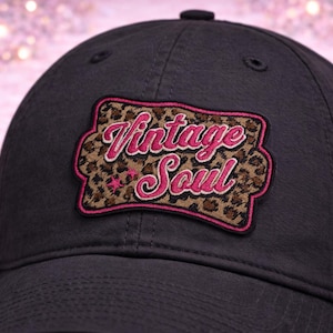 May include: A black baseball cap with a patch that reads "Vintage Soul." The patch has a leopard print background with pink lettering and a pink border. The cap is made of a durable fabric.
