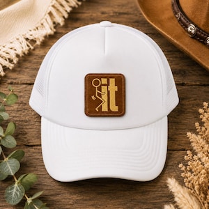 May include: A white trucker hat featuring a square brown patch on the front panel. The patch displays a gold embroidered graphic of a stick figure and the lowercase letters 'it'. The hat has a structured front and a mesh back panel design.