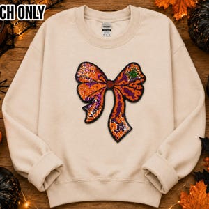 May include: A beige crewneck sweatshirt featuring a large, central graphic of a bow with an orange and purple spiderweb pattern. The text PATCH ONLY is printed in the upper left corner. The sweatshirt has long sleeves and a ribbed collar.