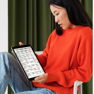 May include: A person wearing an orange sweater and blue jeans holds a tablet displaying a 7-Day Anemia Meal Plan. The tablet screen shows a grid of meal options with text and colorful icons. The background features a green curtain.