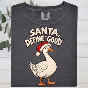May include: A charcoal gray t-shirt featuring a graphic of a white goose wearing a red Santa hat. The text above the goose reads, "SANTA, DEFINE "GOOD"". The shirt has a crew neckline and a Comfort Colors brand label at the inner neck.