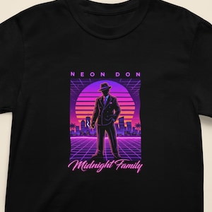 May include: Black t-shirt featuring a retro graphic of a man in a suit and hat, set against a sunset backdrop with the text "NEON DON" and "Midnight Family". The design uses purple, pink, and orange hues.