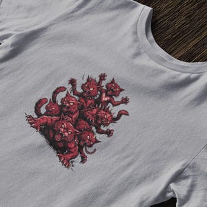 May include: A light gray t-shirt featuring a graphic of several red, cat-like creatures with sharp teeth and claws. The creatures are clustered together, with some in a snarling pose. The design is centered on the shirt.