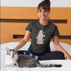 May include: A black t-shirt featuring a stack of five cartoon cats in pastel colors. The cats are pink, yellow, blue, and peach, with hearts and a smiling expression. The shirt is short-sleeved and made of a soft material.