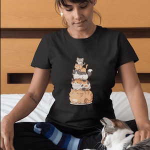 May include: Black t-shirt featuring a stack of cartoon cats in various colors, including orange, gray, and white. The cats are arranged in a playful tower design. The shirt is worn by a person sitting on a bed.