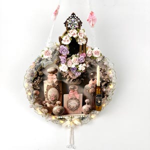 May include: A circular wall hanging featuring a collection of miniature perfume bottles, a candle labeled Witch's Brew, and artificial pink and purple roses. The arrangement is decorated with lace, pearls, and small lights, suspended by white ribbons.