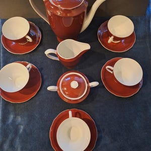 May include: A red and white tea set comprising a teapot, creamer, sugar bowl, and six teacups with saucers. The set has a glossy finish and a classic design, ideal for serving tea.