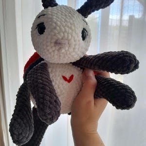 May include: A plush, crocheted stuffed animal featuring a white head and torso with dark gray limbs and antennae. The toy has large black eyes, a small embroidered red heart on its chest, and a bright red shell visible on its back. The texture is soft.
