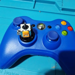 May include: A blue Xbox 360 controller, partially dismantled, with a component exposed. The controller features coloured buttons and a silver central button. The background is a blue surface.
