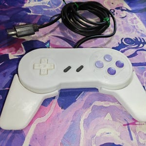 May include: A white retro video game controller with a black cable and connector. The controller has a directional pad, four purple buttons, and two black buttons. The controller is on a colorful, patterned surface.