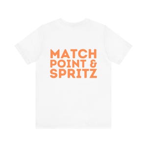 Könnte beinhalten: Weißes T-Shirt mit dem orangefarbenen Text "MATCH POINT & SPRITZ". Der Text ist fett und in einer serifenlosen Schriftart gehalten. Das T-Shirt hat einen Rundhalsausschnitt und kurze Ärmel. Der Text ist mittig auf der Vorderseite des Kleidungsstücks platziert.