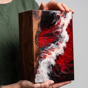 May include: A rectangular wooden box with a striking design. One side features natural wood grain, while the other showcases a vibrant red, white, and black resin wave pattern. The box is held by a person.