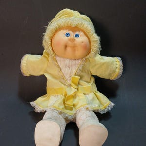 May include: A vintage Cabbage Patch Kid doll with a yellow hooded jacket, dress, and matching bow. The doll has blue eyes, rosy cheeks, and white lace-trimmed socks. The doll's outfit is trimmed with white lace.