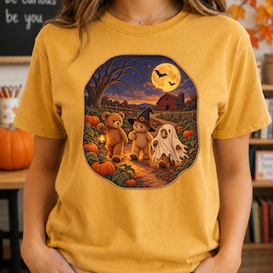May include: A mustard yellow t-shirt featuring a circular graphic of three teddy bears in a pumpkin patch at night. One bear holds a lantern, one wears a witch hat, and one is dressed as a ghost. A red barn and a full moon are in the background.