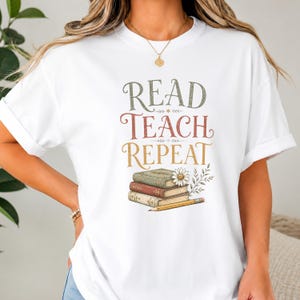 May include: A white short-sleeved t-shirt featuring the text READ TEACH REPEAT in stacked green, red, and gold lettering. Below the text is an illustration of three stacked books, a white daisy flower, and a yellow pencil on a white background.