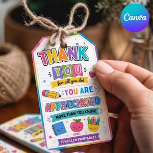 Op de afbeelding: Een kleurrijke cadeaulabel met de tekst "THANK YOU" en "YOU ARE APPRECIATED". De label bevat illustraties van een potlood, appel en notitieboek. Het wordt vastgehouden door een hand en vastgebonden met touw.