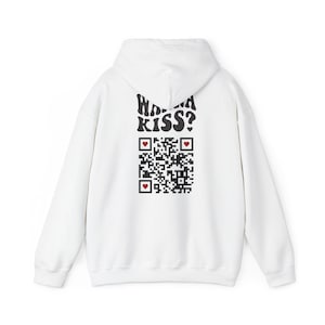Puede incluir: Sudadera con capucha blanca con capucha y mangas largas. La parte trasera de la sudadera presenta el texto "Wanna Kiss?" en negro, en una fuente estilizada sobre un código QR con acentos de corazón rojo. La sudadera está hecha de un material suave.
