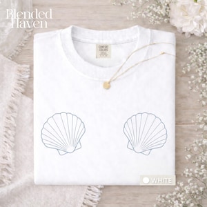 Könnte beinhalten: Weißes T-Shirt mit zwei hellblauen Muschelmotiven. Das Shirt hat einen Rundhalsausschnitt und kurze Ärmel. Eine goldene Halskette mit rundem Anhänger ist über das Shirt drapiert. Das Wort "WHITE" ist auf einem kleinen weißen Etikett aufgedruckt.
