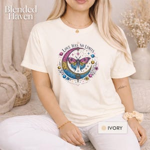 May include: An ivory t-shirt featuring a graphic print of a moth centered on a crescent moon. The design includes the text "LOVE HAS NO LIMITS" and "PANSEXUAL PRIDE" with colorful planets, stars, and floral accents in pink, yellow, and blue.