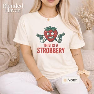May include: Ivory-colored t-shirt featuring a cartoon strawberry with a menacing expression, holding two guns. The text "THIS IS A STROBBERY" is printed below the strawberry in red. The shirt has short sleeves.