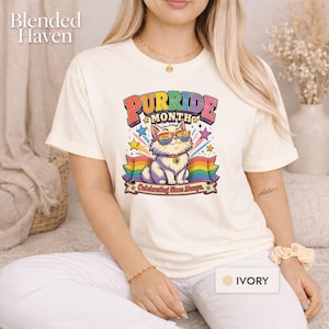 May include: An ivory t-shirt featuring a graphic of a fluffy white cat wearing rainbow-coloured sunglasses. The text reads Purride Month and Celebrating Since Always. The design includes colourful stars, a rainbow banner, and a paw print charm.