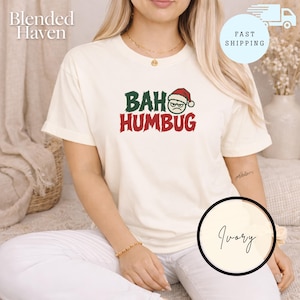 Peut inclure: Un t-shirt à manches courtes ivoire avec le texte BAH HUMBUG en lettres vertes et rouges. La lettre O du mot BAH est remplacée par un visage circulaire vert avec une expression grincheuse portant un bonnet de Père Noël rouge et blanc.