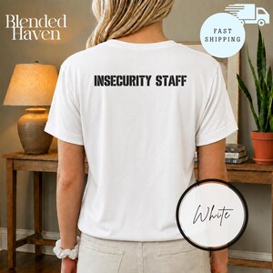 Op de afbeelding: Een wit T-shirt met korte mouwen en de tekst INSECURITY STAFF in zwarte blokletters op de bovenrug. Het T-shirt heeft een ronde hals en een standaard pasvorm, weergegeven vanaf de achterkant op een persoon.